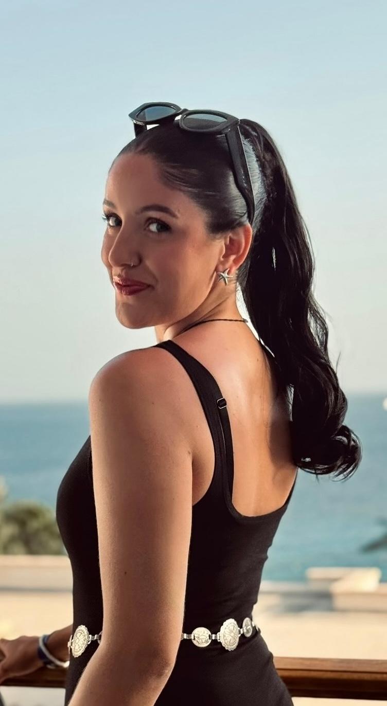 Kvinna med långt mörk tofs, svart t-shirt och solglasögon på huvudet som ler med havet bakom sig.