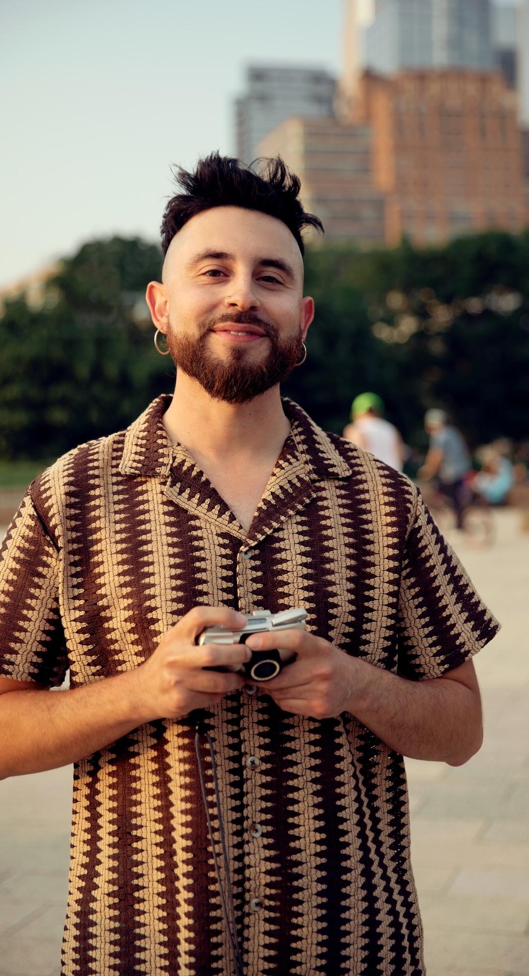 Skäggig man i mönstrad skjorta som håller en kamera och står utomhus med träd och byggnader i bakgrunden.