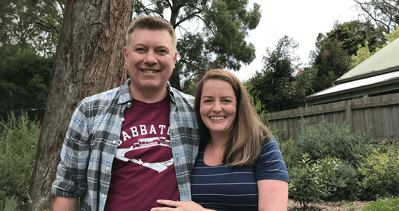 Jude and Ben are standing together outside. Ben is wearing a plaid shirt over a red t-shirt and Jude is wearing a blue top. Both are smiling happily.